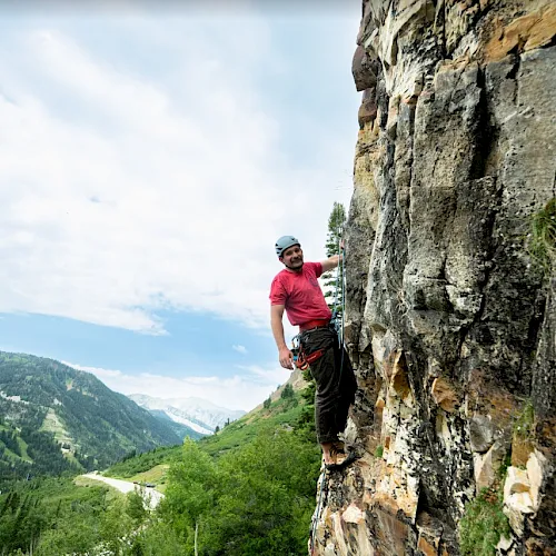 A mountain climber in a red shirt scales a rugged rock face above a lush valley and distant peaks under a blue sky.