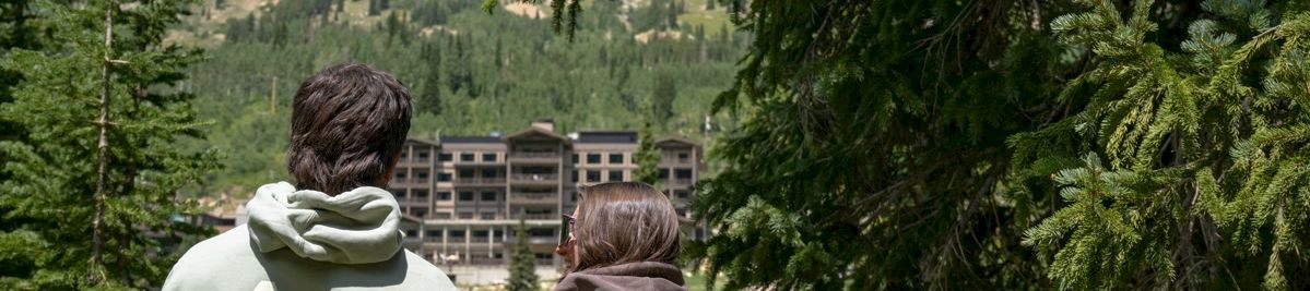 Two people in hoodies stand watching a mountain valley and hotel buildings through pine trees.