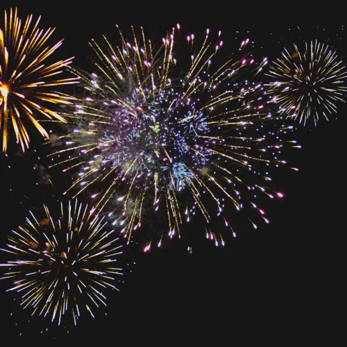 Colorful fireworks burst across a dark night sky, with gold, blue, and pink trails lighting up the scene.
