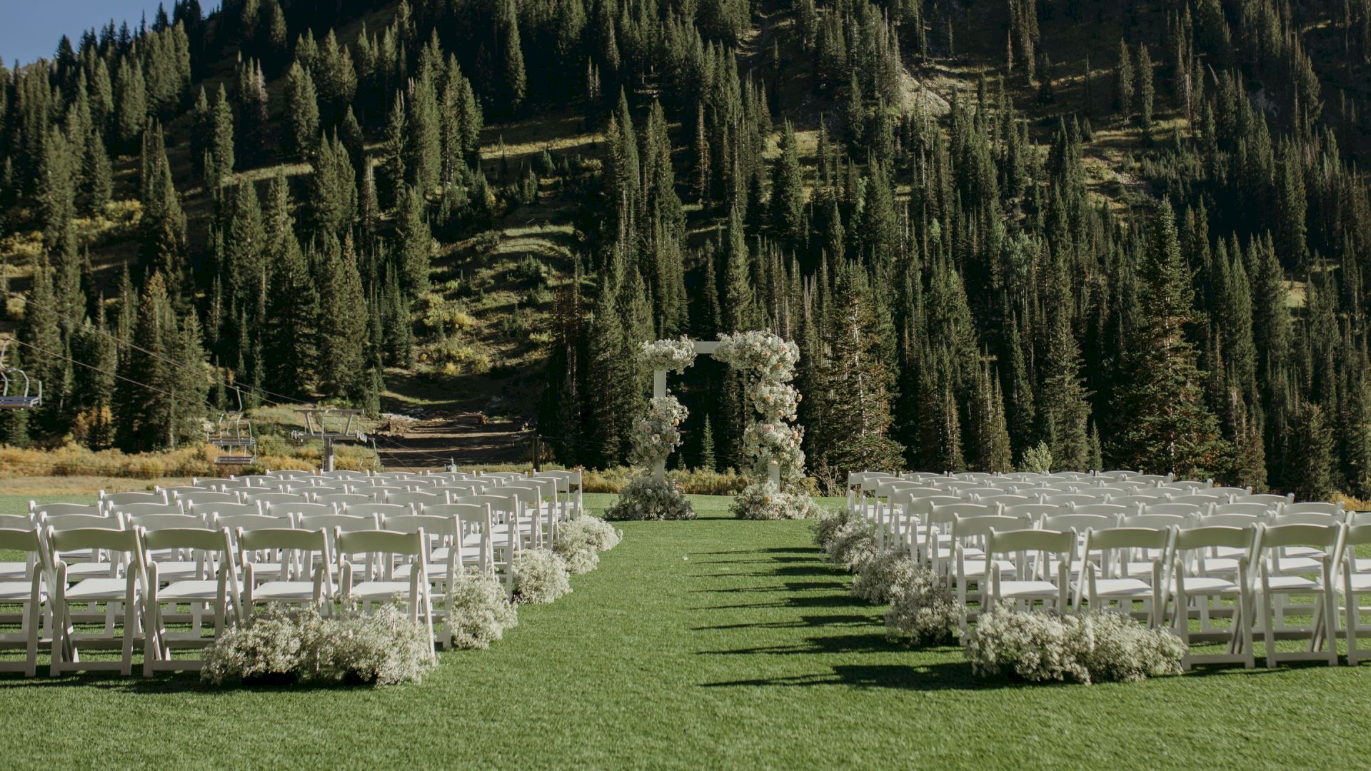 Outdoor wedding setup with white chairs, a floral arch, green lawn, and pine-covered mountain backdrop under a clear sky.
