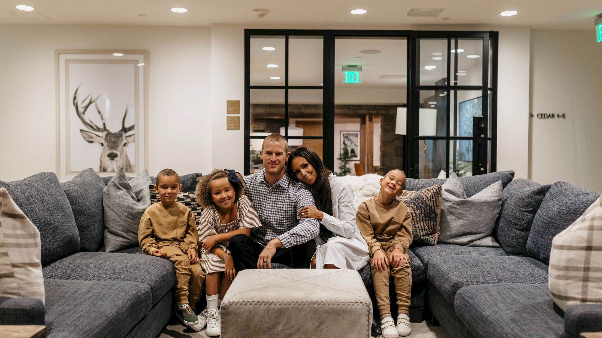 A family of five poses on a cozy gray sofa in a bright living room with modern glass doors behind them.