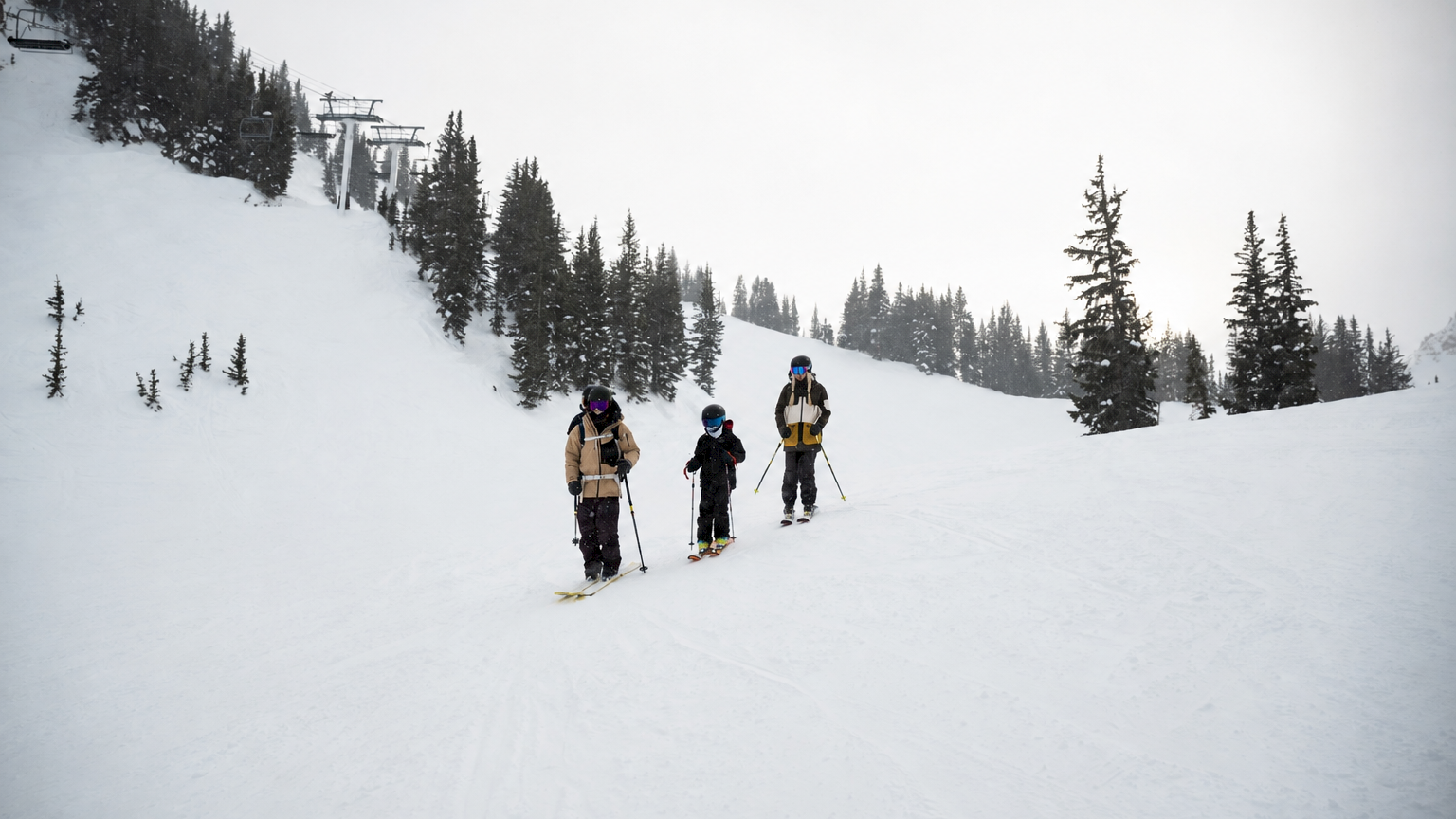 Three skiers hike down a snowy slope near a chairlift, surrounded by pine trees in overcast mountains.