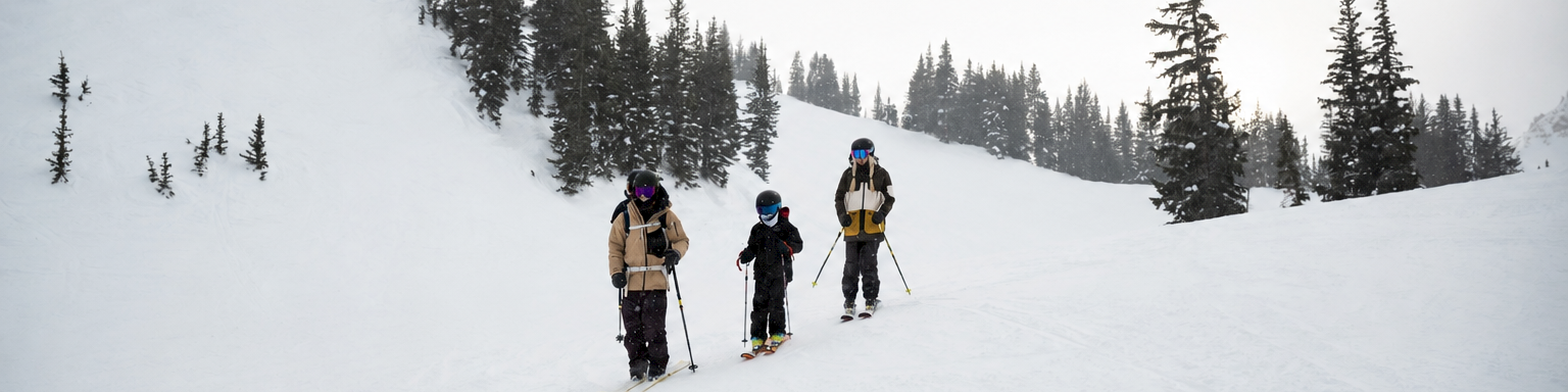 Three skiers hike down a snowy slope near a chairlift, surrounded by pine trees in overcast mountains.