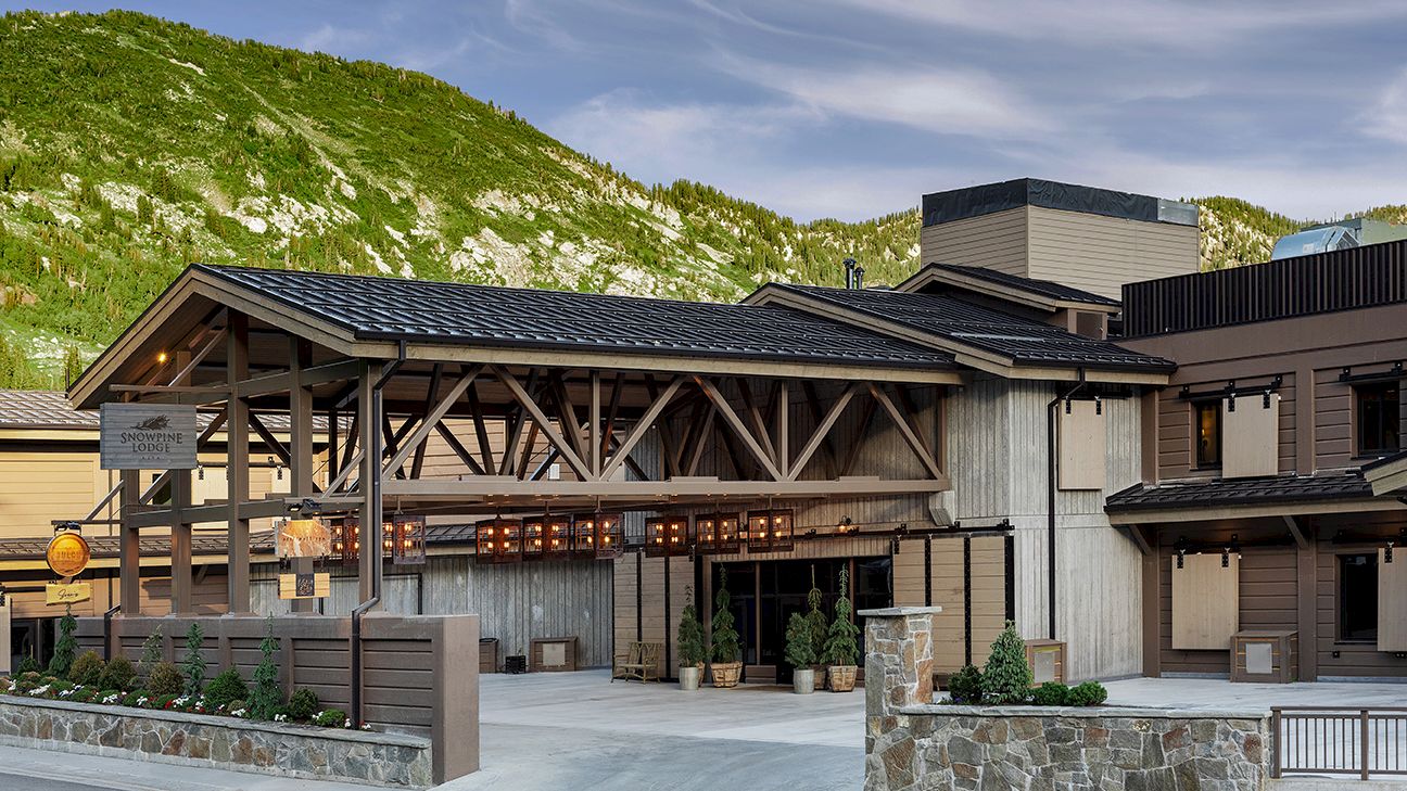 A mountain lodge building with stone base, dark roofs, and landscaped plants against a green hillside.