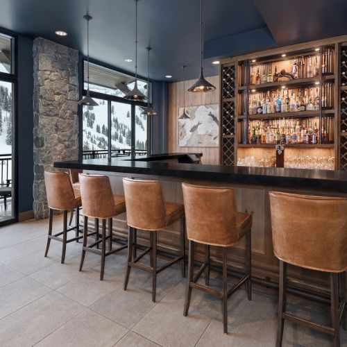 A cozy mountain bar with a bottle display, leather stools, pendant lights, and large windows showing snowy pine views.