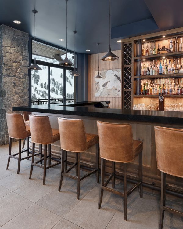 A cozy mountain bar with a bottle display, leather stools, pendant lights, and large windows showing snowy pine views.