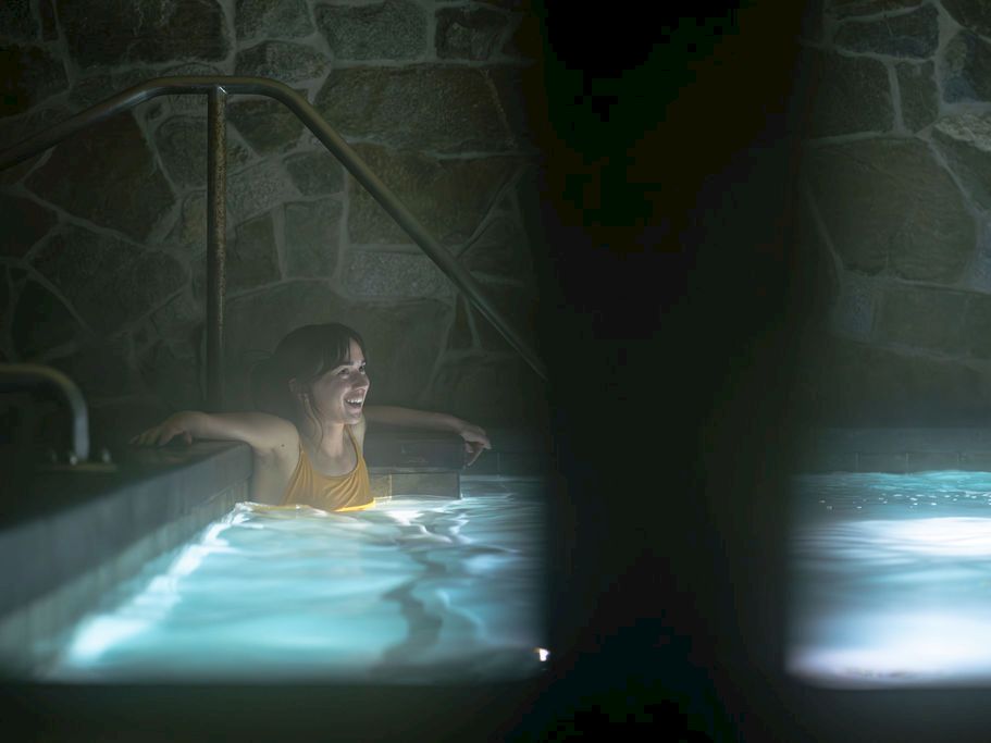 A woman in a yellow top relaxes in an indoor pool, seen through dark foreground shadows and a stone wall.