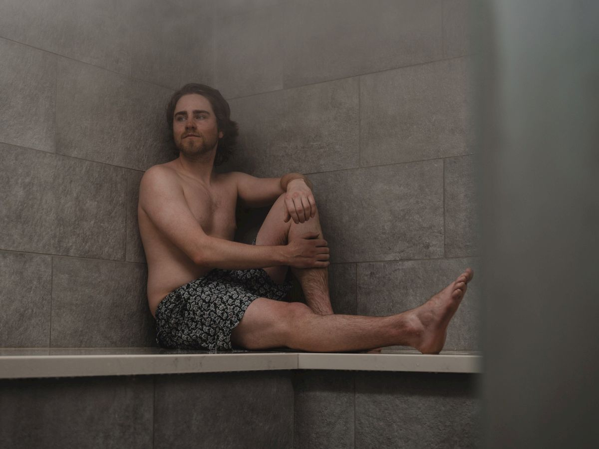A shirtless man lounges in a tiled bathtub, legs outstretched and relaxed, looking toward the camera in a bathroom.