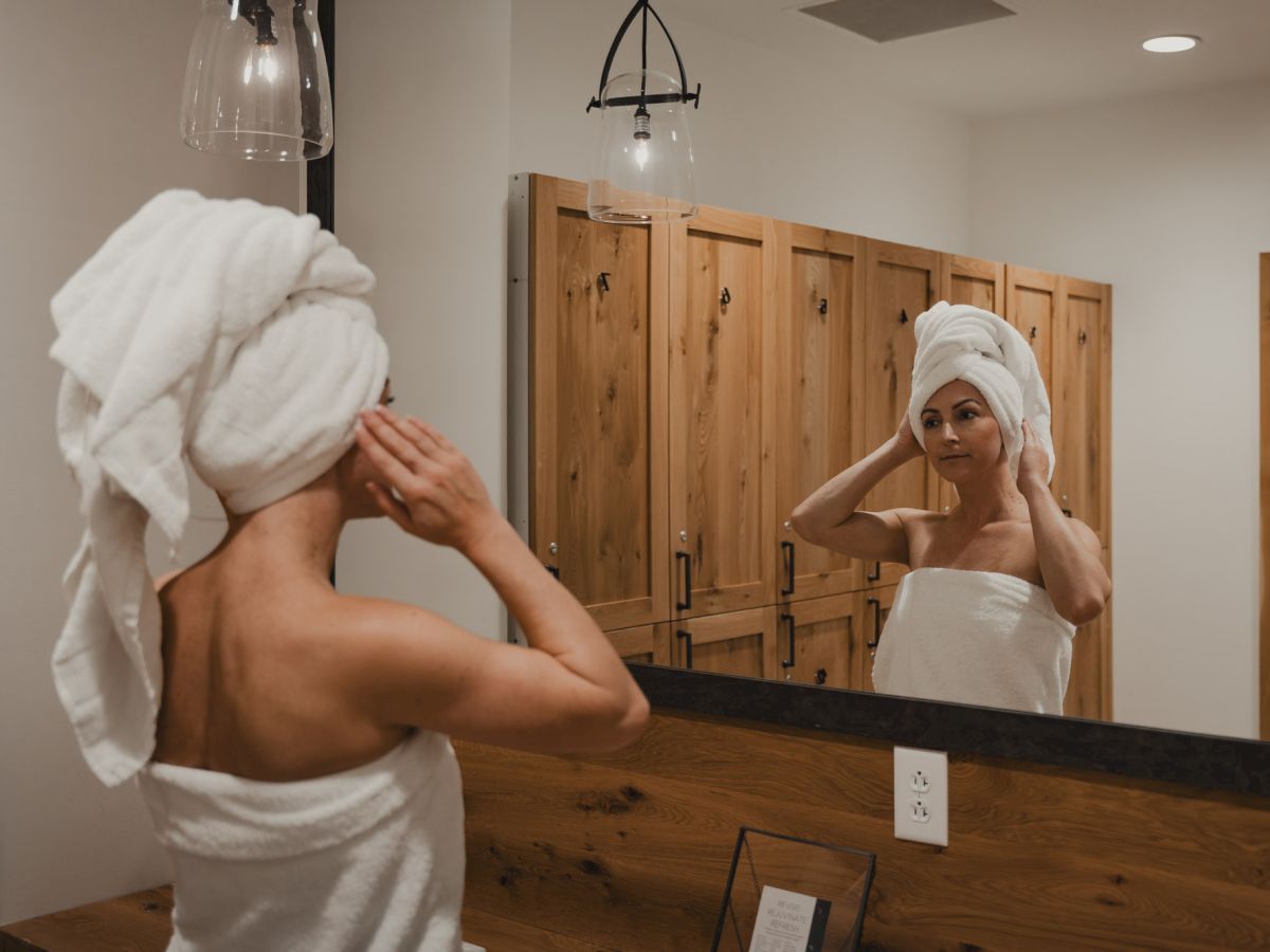 A woman in a white robe adjusts her towel in a bathroom mirror, seen from behind with wooden cabinets behind her.