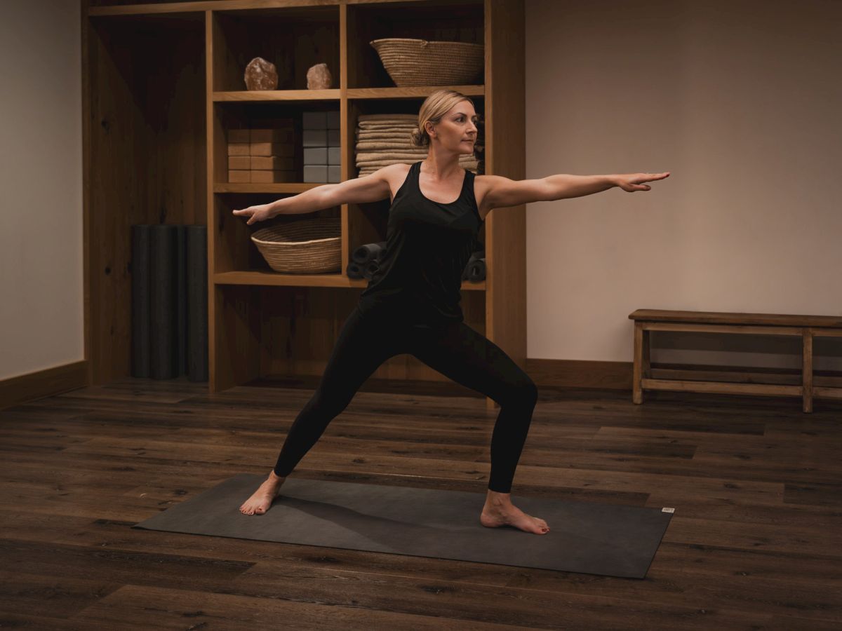A woman in black doing a yoga lunge/stretch in a warm studio, arms extended on a mat.