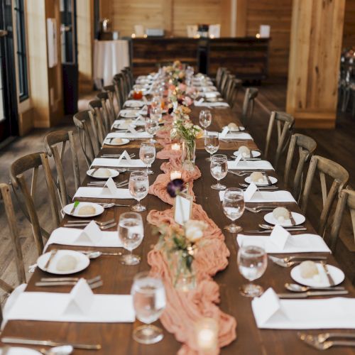 A rustic barn dinner setup with long tables, white plates, candles, glasses, and pink floral centerpieces for a celebration.