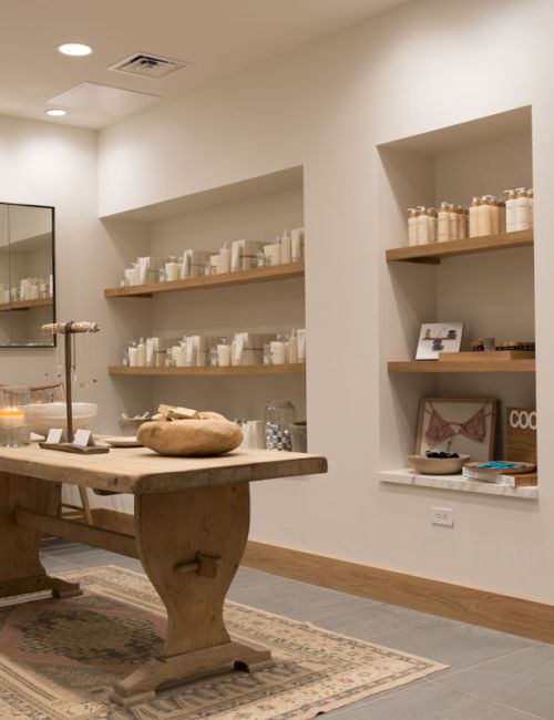 A bright boutique spa room with a wooden treatment table, candles, and neatly stocked shelves of toiletries.