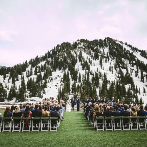 Outdoor wedding ceremony on a grassy lawn with guests seated, facing a couple at a snowy mountain peak under overcast skies.