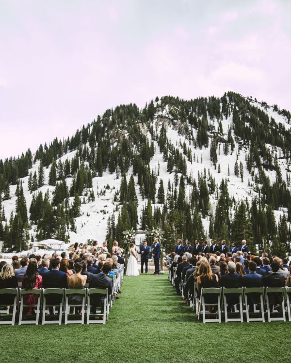 Outdoor wedding ceremony on a grassy lawn with guests seated, facing a couple at a snowy mountain peak under overcast skies.