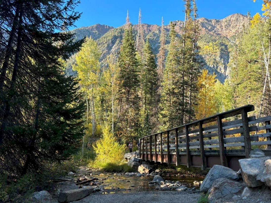 Scenic mountain forest with a wooden bridge over a shallow stream, golden autumn trees, and distant hikers.