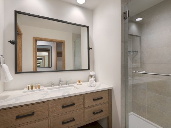 A modern bathroom with a double vanity, large framed mirror, toiletries, and a glass-walled shower with towel bars.