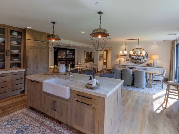 A modern kitchen and dining area with a stone island sink, pendant lights, wood cabinetry, and a living room beyond.