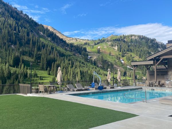 An outdoor resort pool and patio with loungers, set against lush alpine mountains under a blue sky.