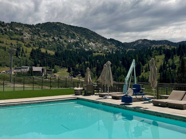 An outdoor pool and patio with lounge chairs and umbrellas overlooks a lush green valley and steep mountain slopes.