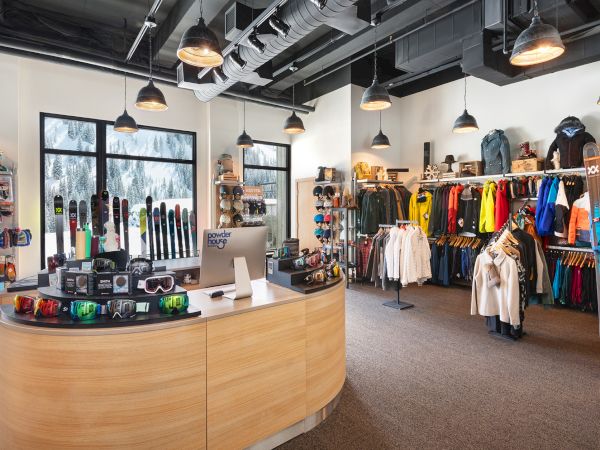 A modern ski shop interior filled with snowboards, skis, goggles, helmets, and colorful winter jackets hanging on racks under warm lights.