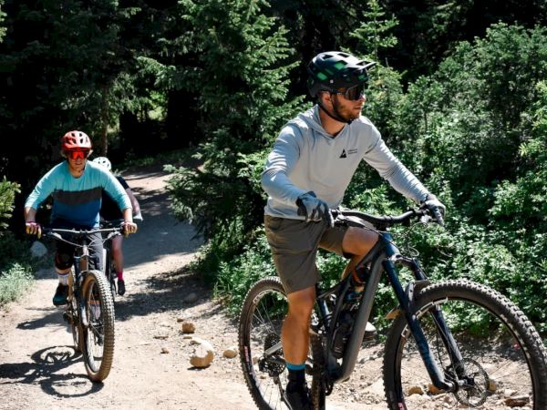 Two cyclists in helmets ride mountain bikes on a sunny forest trail, navigating a rocky path.