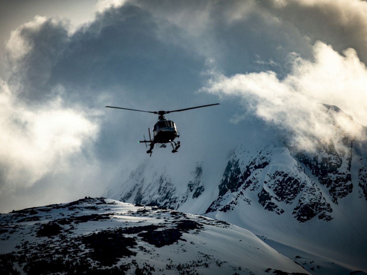A helicopter flies over snowy mountain peaks as clouds swirl around it.