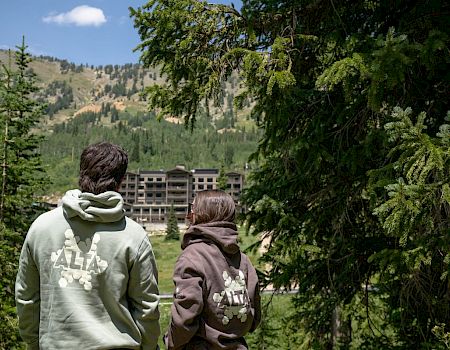Two people in hoodies stand watching a mountain valley and hotel buildings through pine trees.