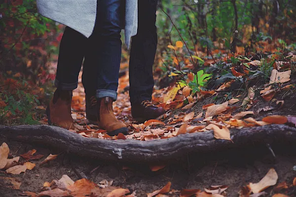A person&rsquo;s legs in jeans and brown boots walk through a forest floor scattered with colorful autumn leaves, embracing fall.