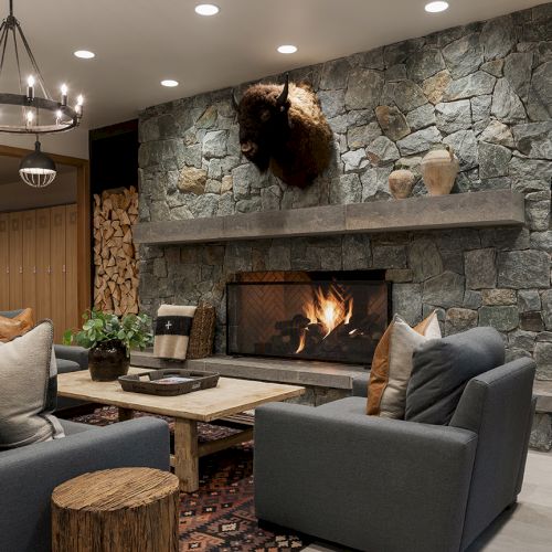 A cozy living room with gray sofas, a large stone fireplace with fire, and a hanging buffalo head above the mantel.