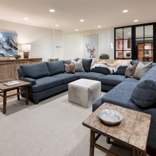 A modern living room with a blue sectional sofa, deer-themed art, wood sideboards, coffee tables, and large black-framed glass doors.