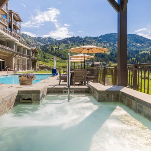 Relax in a heated hot tub on a balcony, overlooking a pool, umbrellas, and green mountain valleys under a blue sky.