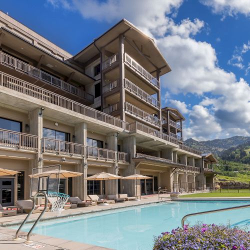 A luxury hotel terrace with a blue swimming pool, patio umbrellas, and stacked balcony rooms beneath a bright mountain sky.