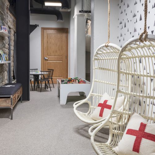 A cozy kids playroom with a white woven rocking chair, bookshelves, toys, and a patterned wall in a stone hallway.