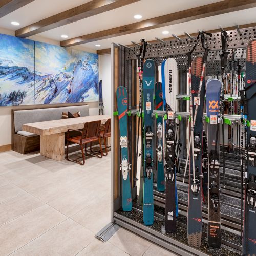 A ski storage room packed with colorful skis on a wall, with a bench and snowy mountain mural in the background.