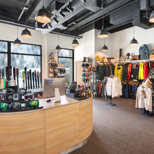 A brightly lit ski shop filled with skis, goggles, helmets, and colorful winter jackets by large windows and hanging lights.