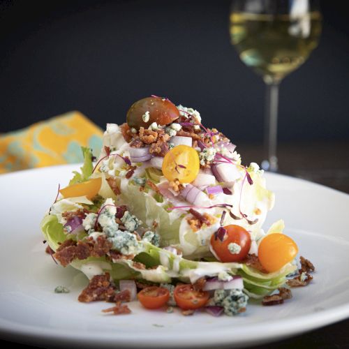 A gourmet salad piled with greens, cherry tomatoes, blue cheese, bacon, and herbs, with a wine glass blurred in back.
