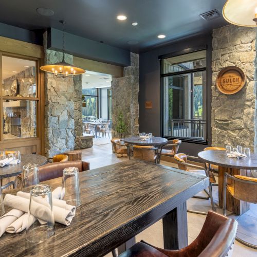 Cozy restaurant dining area with wooden tables, stone pillars, pendant lights, and big windows showing a forested mountain view.