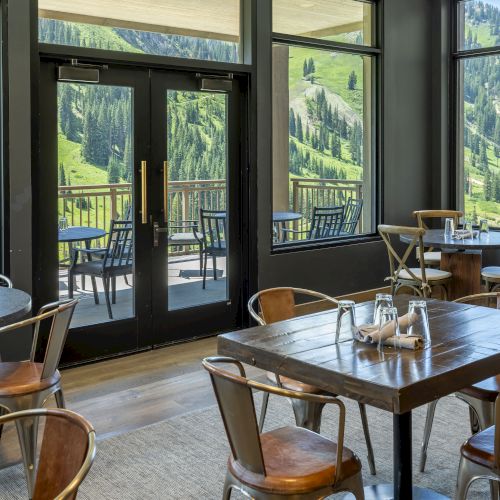 A cozy restaurant dining area with wooden tables and chairs, framed by large windows overlooking lush green mountains and forests.