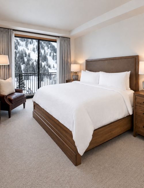 A cozy hotel bedroom with a large bed, side lamps, and curtains framing snowy pine-covered mountains outside.