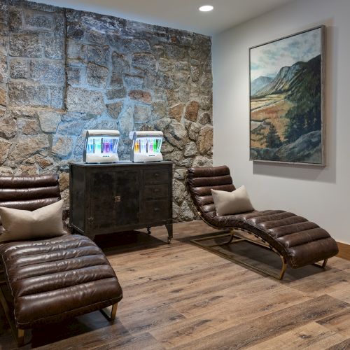 A spa-like room with brown recliners, a stone wall, a cabinet holding twin light-therapy machines, and a landscape painting.