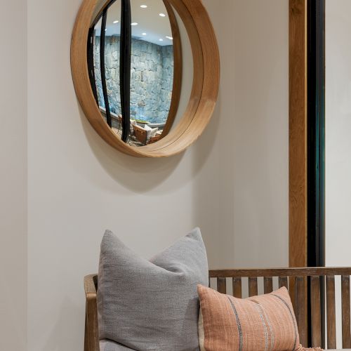 An indoor lounge shows a wooden bench with throw pillows, a round wooden-framed mirror, and a reflected stone wall across windows.