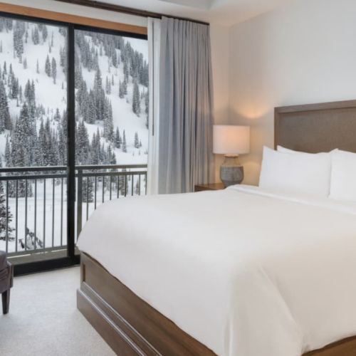A cozy hotel bedroom with a large bed, bedside lamps, and gray curtains framing a snowy forest view from the balcony.