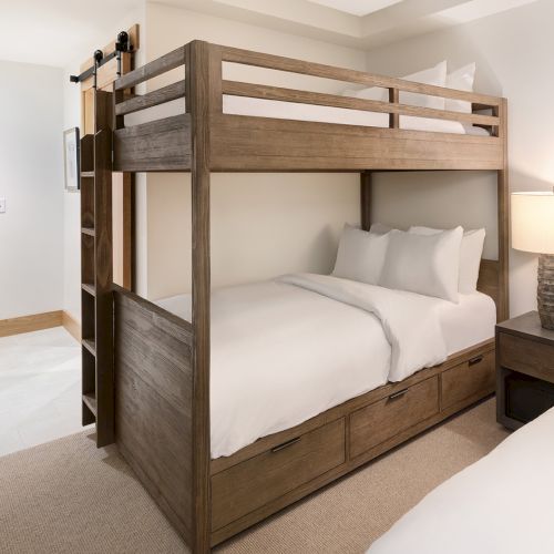 A modern bedroom shows a wooden bunk-style platform bed with open shelves, neatly made white sheets, and bedside lamps.