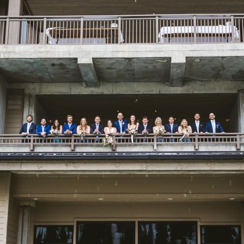 A wedding party poses on a building balcony in blue suits and dresses, holding bouquets and wearing boutonnieres.