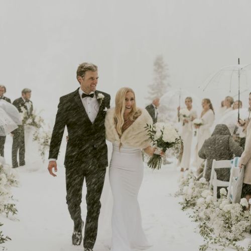 A snowy outdoor wedding ceremony: a tuxedoed groom and bride in white walk down an aisle as guests hold umbrellas.