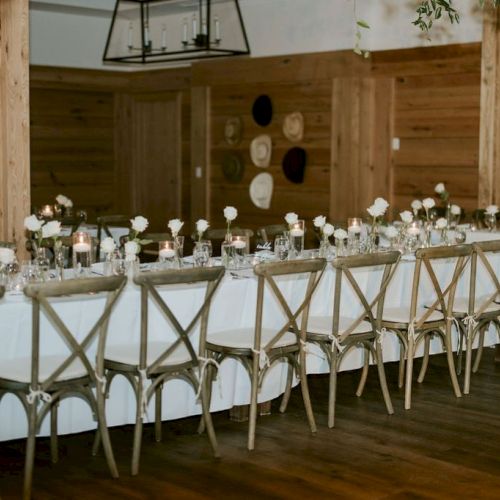 Wedding-style banquet setup with a long table, gold chairs, candles, and white flowers in a rustic wooden hall.