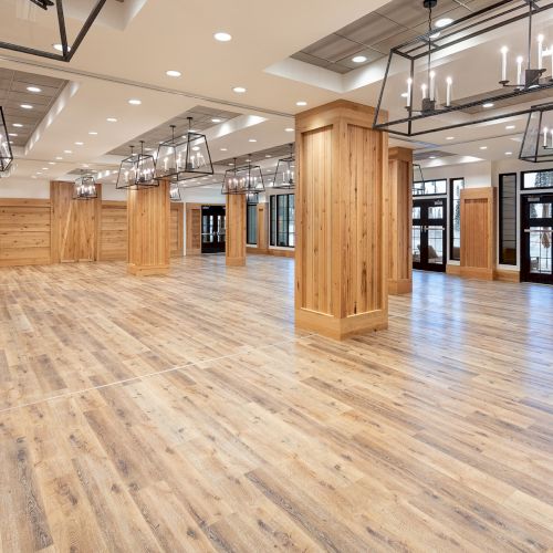 Bright modern event hall with warm wood paneling, polished floor, and hanging black chandeliers overhead.