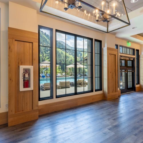 Indoor hallway/lobby with wood paneling, large windows showing a pool and forested mountains, plus hanging lights.