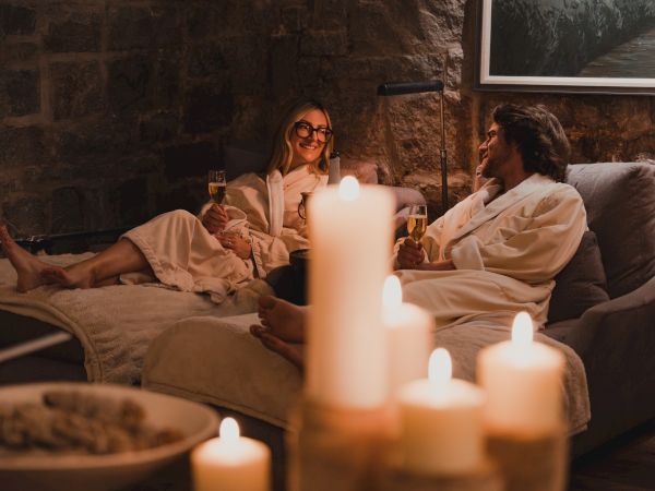 A cozy couple in bathrobes reclines on a couch, smiling with champagne flutes by candlelight and a warm fireplace glow.