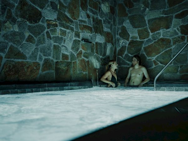 Two people relax in a glowing indoor hot tub, splashing water from a wall feature against a tiled stone backdrop.